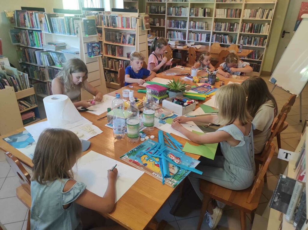 na zdjęciu widać salę biblioteczną, przy stole siedzą dzieci, wykonują prace plastyczne