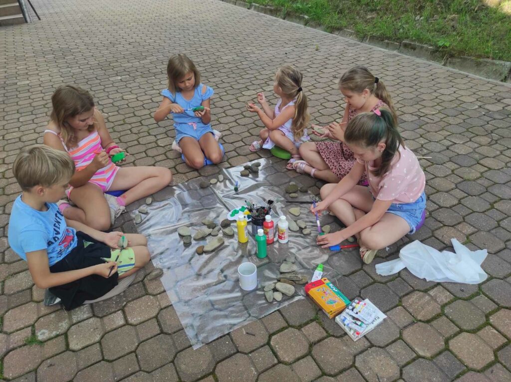 na zdjęciu widać fragment podwórka przed biblioteką, na ziemi rozłożone są materiały plastyczne, dzieci siedzą wokół i wykonują prace plastyczne