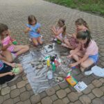 na zdjęciu widać fragment podwórka przed biblioteką, na ziemi rozłożone są materiały plastyczne, dzieci siedzą wokół i wykonują prace plastyczne