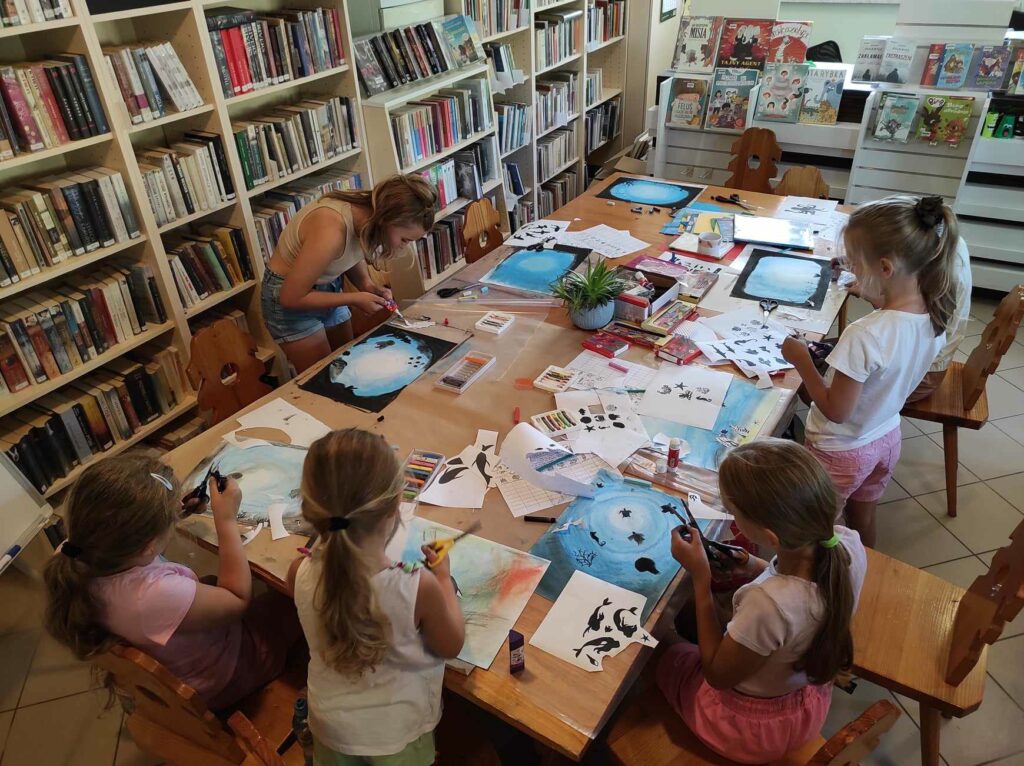 na zdjęciu widać salę biblioteczną, przy stole siedzą dzieci