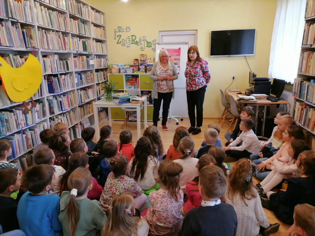 Na zdjęciu widoczna jest autorka książek, która wraz z pracownikiem biblioteki wita wszystkie dzieci przybyłe na spotkanie. Dzieci siedzą na podłodze na kolorowych poduszkach. W tle regały z książkami, regał z grami planszowymi, telewizor i biurko z komputerami oraz stolik na którym stoi wazon z kwiatami i położone są książki autorki.