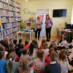Na zdjęciu widoczna jest autorka książek, która wraz z pracownikiem biblioteki wita wszystkie dzieci przybyłe na spotkanie. Dzieci siedzą na podłodze na kolorowych poduszkach. W tle regały z książkami, regał z grami planszowymi, telewizor i biurko z komputerami oraz stolik na którym stoi wazon z kwiatami i położone są książki autorki.