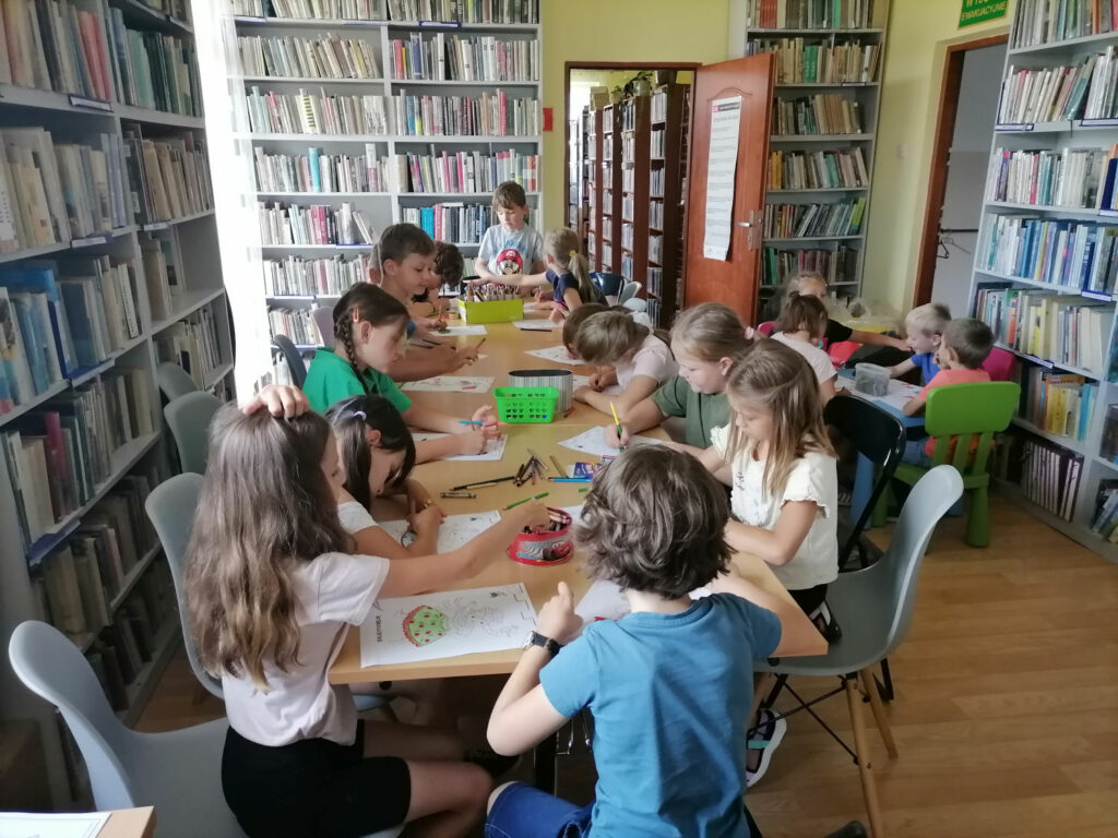 Na zdjęciu widoczna jest grupa dzieci siedzących przy stolikach i kolorujących stroje ludowe. W tle regały z książkami.