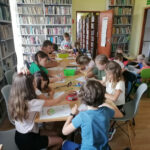 Na zdjęciu widoczna jest grupa dzieci siedzących przy stolikach i kolorujących stroje ludowe. W tle regały z książkami.
