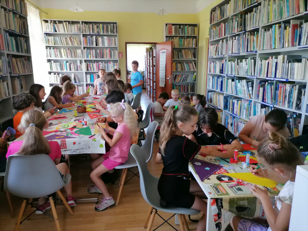 Na zdjęciu widoczna jest grupa dzieci siedzących przy stolikach i wykonujących chusty z kolorowego papieru i włóczki oraz góralskie kapelusze z papieru piórek i muszelek. W tle regały z książkami.