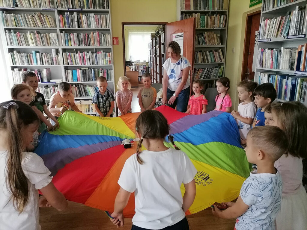 Na zdjęciu widoczna jest grupa przedszkolaków wraz z wychowawcą stojących wokół chusty animacyjnej i podrzucających na niej maskotkę Pucia.
