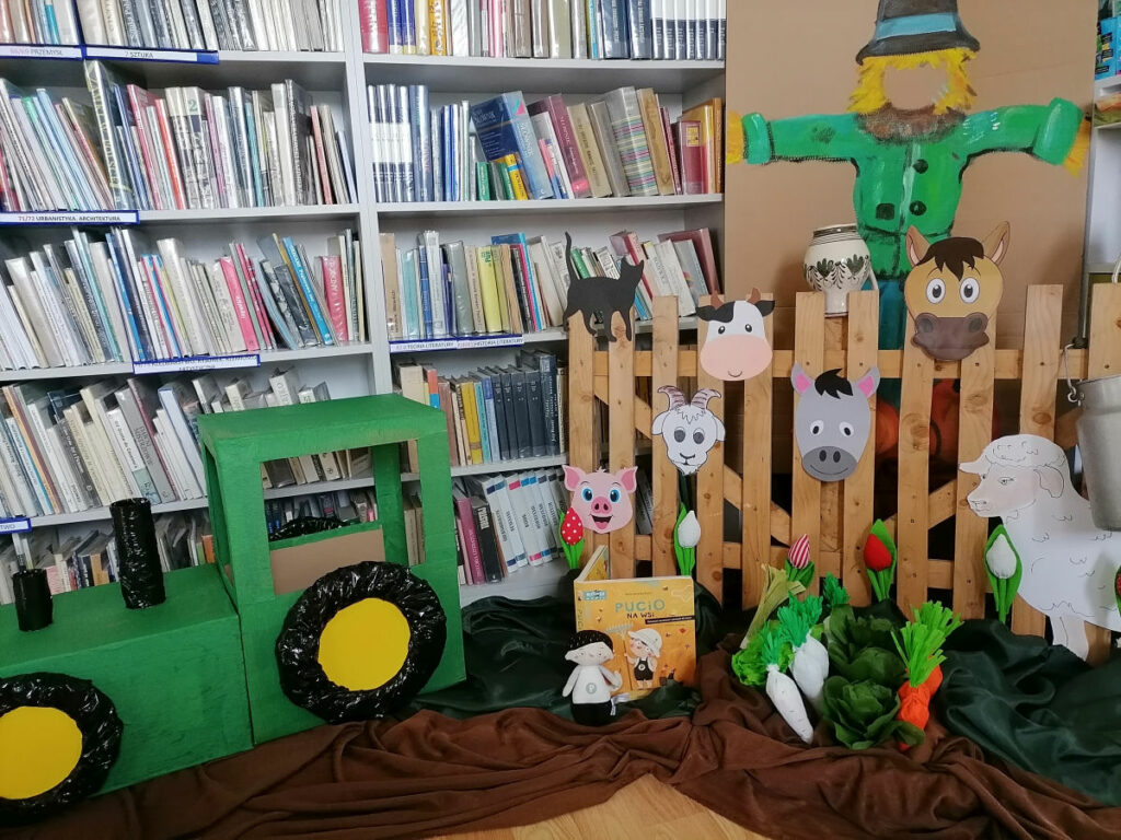 a zdjęciu widoczna jest dekoracja wykorzystana do zabawa podczas zajęć są to: tekturowy traktor, strach na wróble płotek z głowami zwierząt, papierowy warzywa, maskotka Pucia i książka Pucio na wsi.