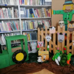 a zdjęciu widoczna jest dekoracja wykorzystana do zabawa podczas zajęć są to: tekturowy traktor, strach na wróble płotek z głowami zwierząt, papierowy warzywa, maskotka Pucia i książka Pucio na wsi.