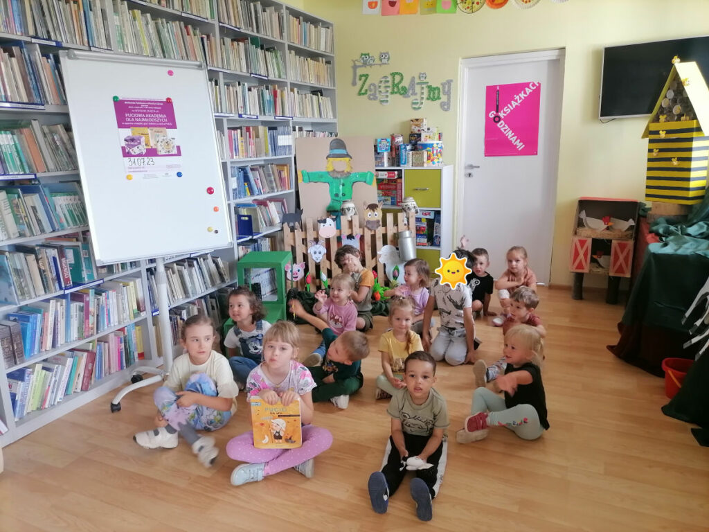 Na zdjęciu widoczna jest grupa przedszkolaków pozujących na tle wiejskich dekoracji. Dzieci siedzą na podłodze, jedna dziewczynka trzyma książkę Pucio na wsi.