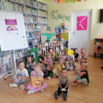Na zdjęciu widoczna jest grupa przedszkolaków pozujących na tle wiejskich dekoracji. Dzieci siedzą na podłodze, jedna dziewczynka trzyma książkę Pucio na wsi.