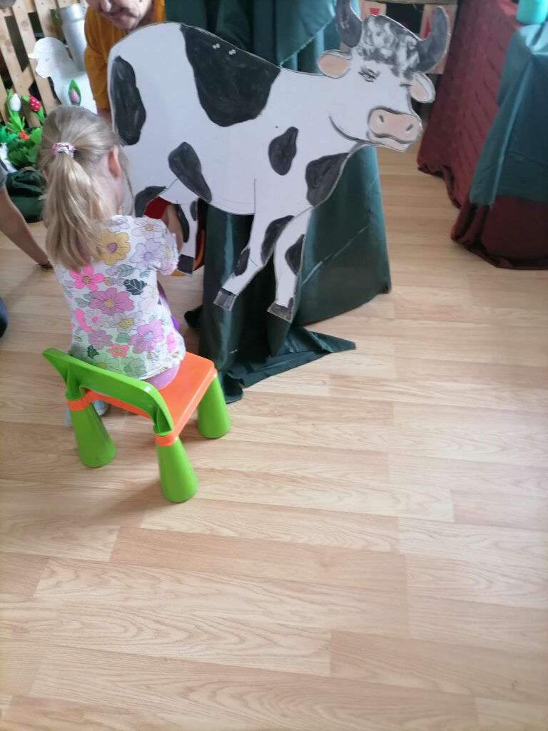 Na zdjęciu widoczna jest dziewczynka, która doi tekturową krowę.