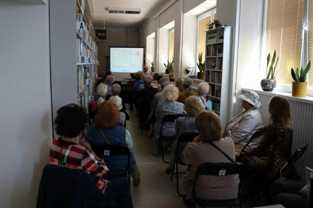 Wnętrze biblioteki centralnej. Na krzesłach podczas spotkania autorskiego siedzą przybyli czytelnicy. Z samego przodu autor spotkania a za nim ekran na którym wyświetlano pokaz slajdów.