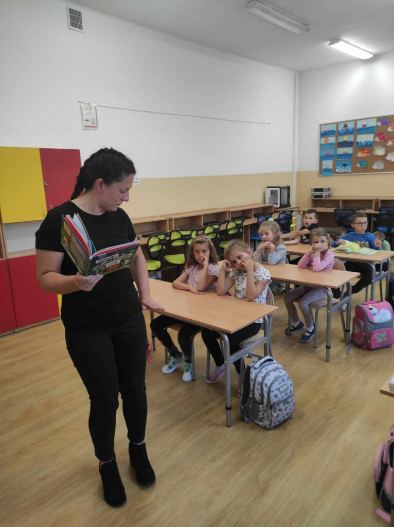 na zdjęciu widać salę szkolną, przy stolikach siedzą dzieci, bibliotekarz przedstawiam dzieciom książkę