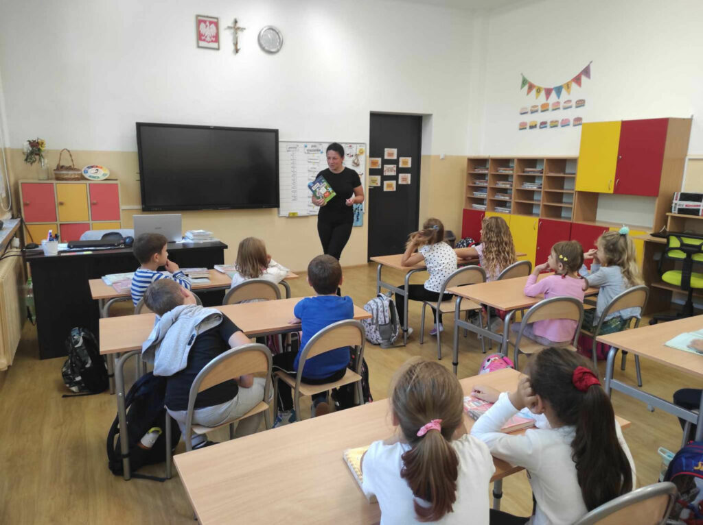 na zdjęciu widać salę szkolną, przy stolikach siedzą dzieci, bibliotekarz przedstawiam dzieciom książkę