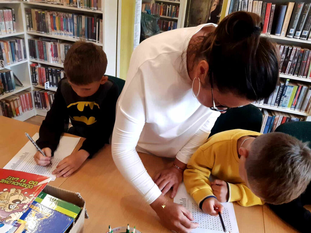 na zdjęciu widać bibliotekarkę oraz chłopców piszących na kartkach w sali bibliotecznej