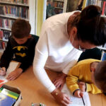 na zdjęciu widać bibliotekarkę oraz chłopców piszących na kartkach w sali bibliotecznej