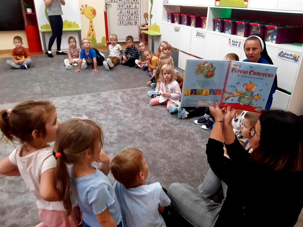 na zdjęciu widać dzieci siedzące na podłodze w sali przedszkolnej i czytającą im bibliotekarkę.