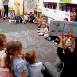 na zdjęciu widać dzieci siedzące na podłodze w sali przedszkolnej i czytającą im bibliotekarkę.