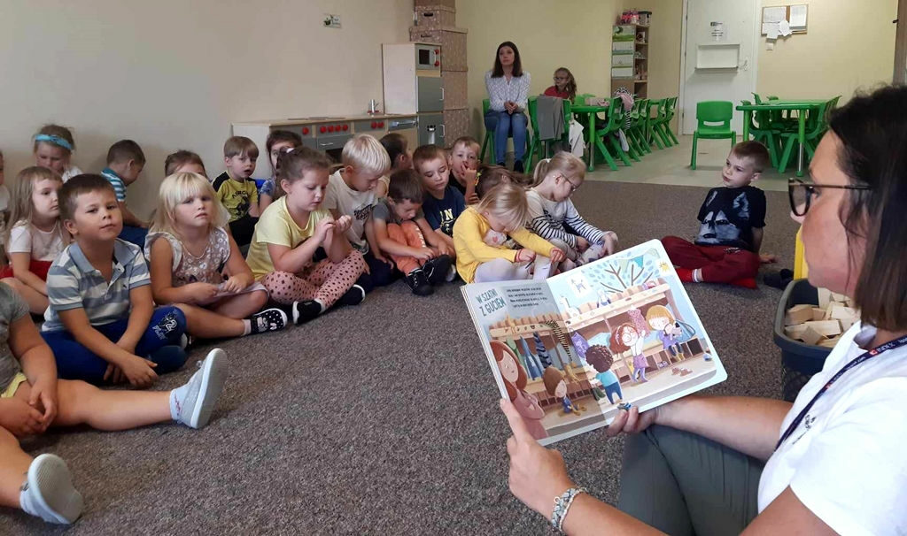 na zdjęciu widać dzieci siedzące na podłodze i słuchające czytającej im bibliotekarki