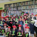 Na zdjęciu widoczna jest grupa 21 dzieci stojących pod regałami z książkami i prezentujących wykonane przez siebie sombrera i kaktusy.