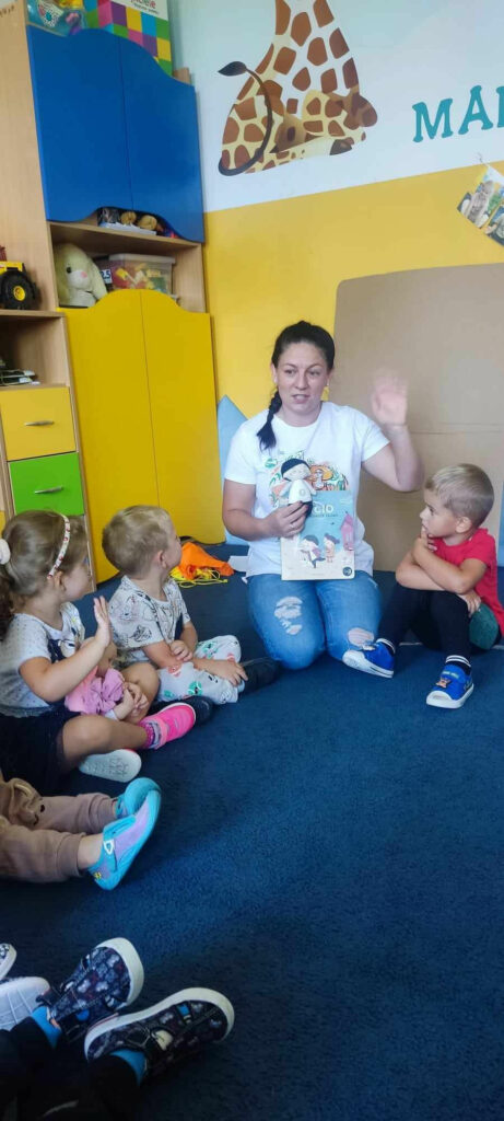 na zdjęciu widać salę przedszkolną, dzieci siedzą na dywanie, bibliotekarz przedstawia bohatera z książki