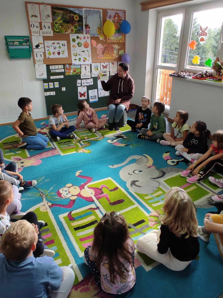 na zdjęciu widać salę przedszkolną, dzieci siedzą w kole na dywanie, bibliotekarz czyta książkę