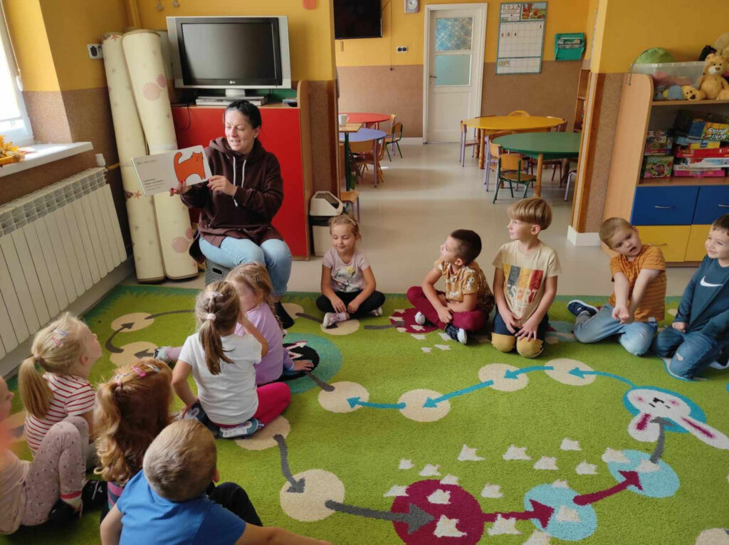 na zdjęciu widać salę przedszkolną, dzieci siedzą w kole na dywanie, bibliotekarz czyta książkę