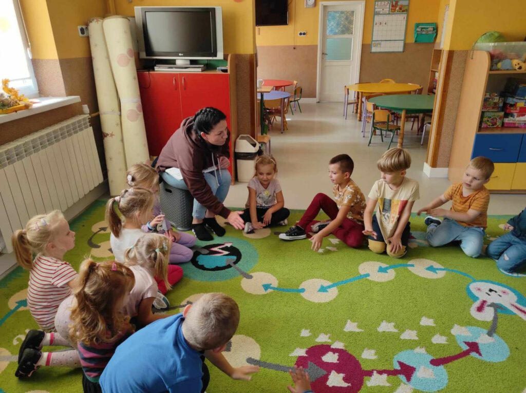 na zdjęciu widać salę przedszkolną, dzieci siedzą w kole na dywanie, bibliotekarz czyta książkę
