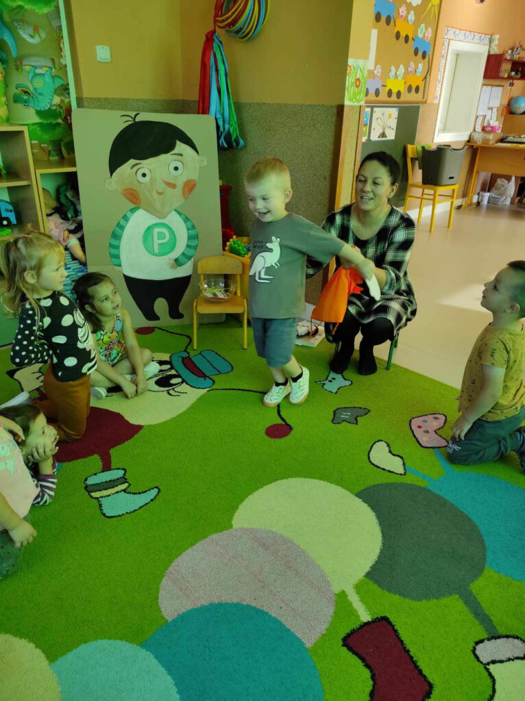 na zdjęciu widać salę przedszkolną, dzieci siedzą na dywanie, bibliotekarz bawi się z dziećmi