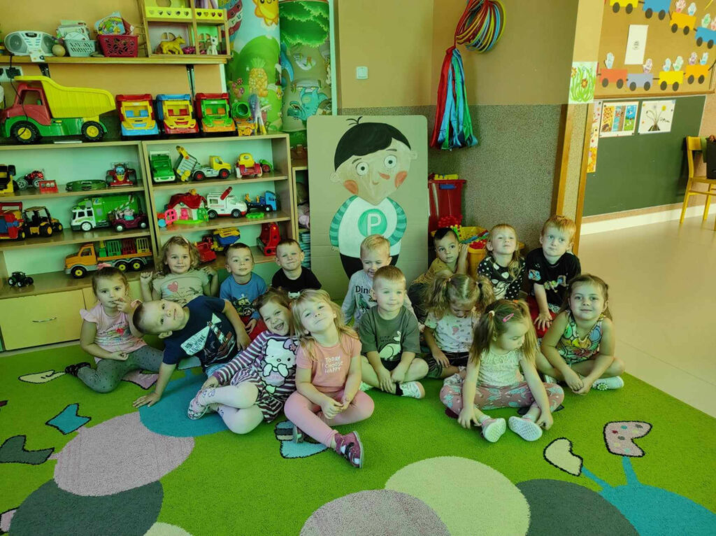 na zdjęciu widać salę przedszkolną, dzieci siedzą na dywanie, w tle widać bohatera czytanej książki - Pucia