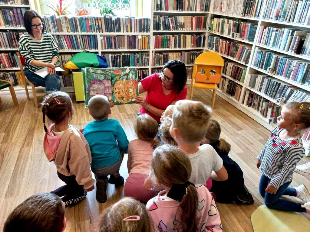 na zdjęciu widać dzieci siedzące na podłodze oraz czytające im bibliotekarki