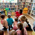 na zdjęciu widać dzieci siedzące na podłodze oraz czytające im bibliotekarki