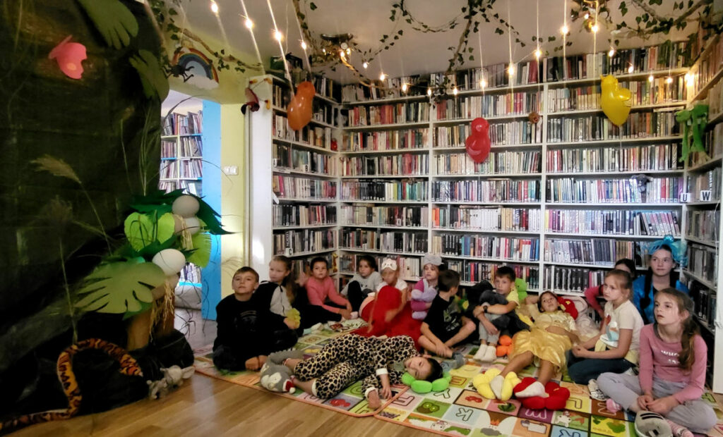 na zdjęciu widać dzieci siedzące w sali bibliotecznej na tle regałów z książkami. Sala udekorowana jest w stylu dżungli.