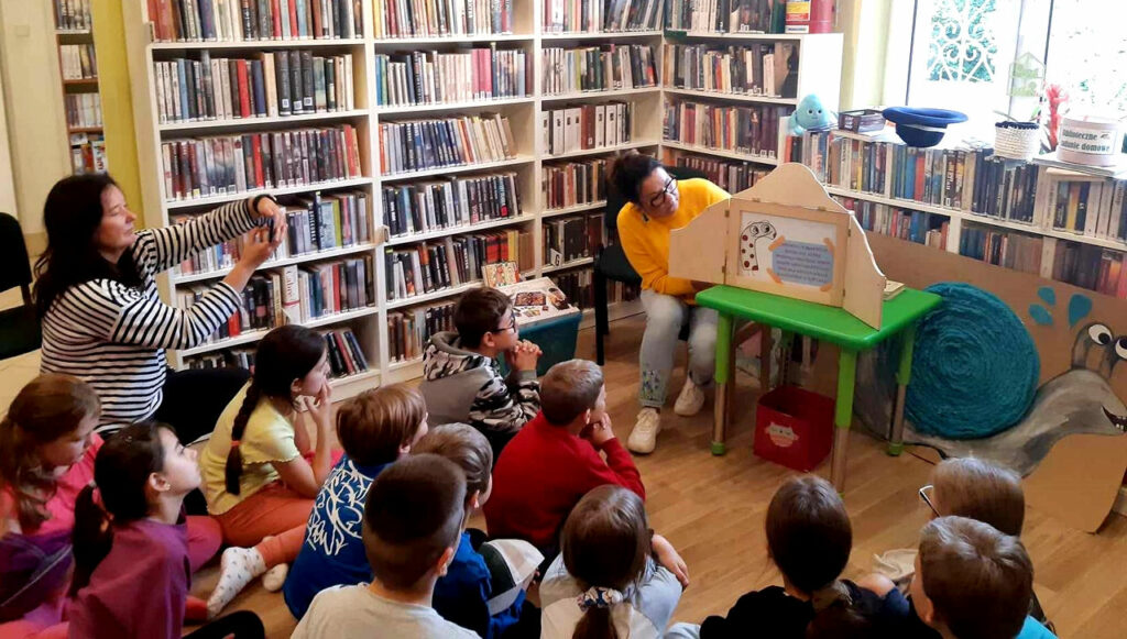 na zdjęciu widać salę biblioteczna, dzieci siedzące na podłodze i czytająca im bibliotekarkę
