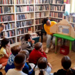 na zdjęciu widać salę biblioteczna, dzieci siedzące na podłodze i czytająca im bibliotekarkę