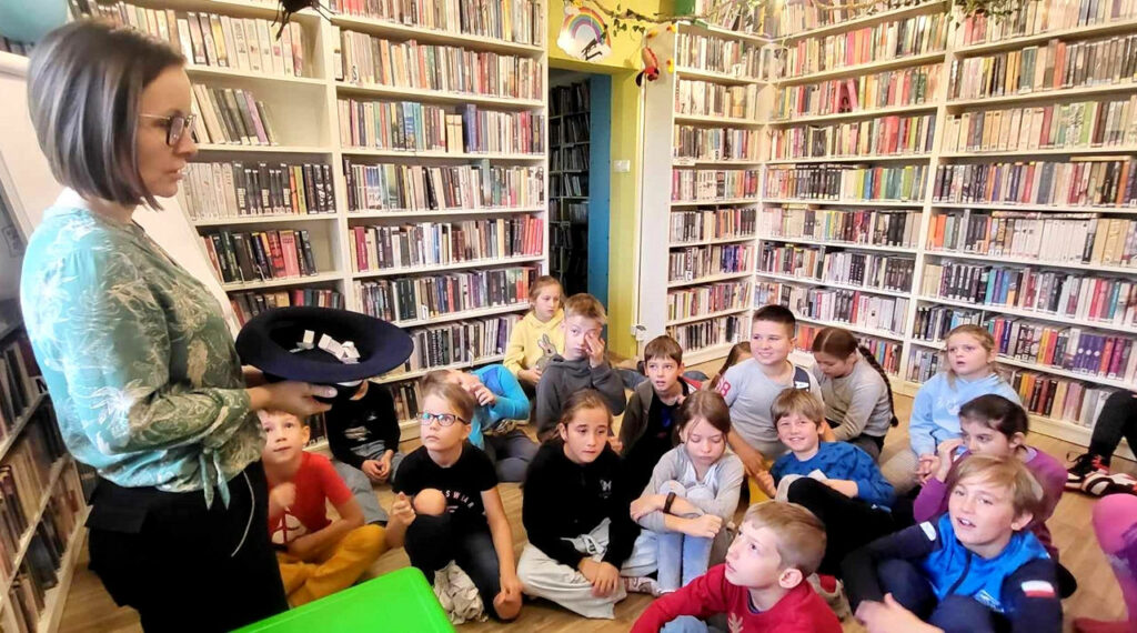 na zdjęciu widać dzieci siedzące na podłodze oraz stojąca przed nimi bibliotekarkę