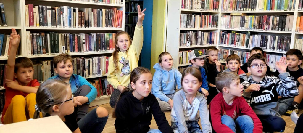 na zdjęciu widać dzieci siedzące na podłodze. W tle widać regały z książkami.