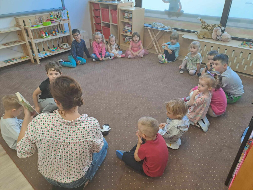 dzieci z przedszkola Montessori siedzą na dywanie i słuchają bajki czytanej przez bibliotekarza
