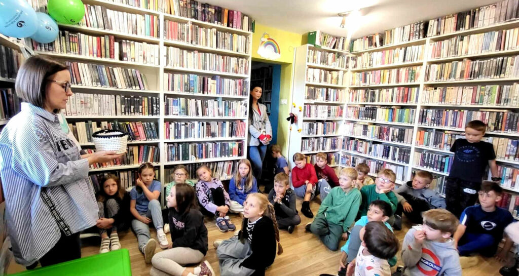 na zdjęciu widać dzieci siedzące na podłodze w sali bibliotecznej i bibliotekarkę trzymająca w ręku pudełko.