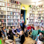 na zdjęciu widać dzieci siedzące na podłodze w sali bibliotecznej i bibliotekarkę trzymająca w ręku pudełko.