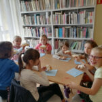 na zdjęciu widoczna jest grupa 7 dziewczynek siedzących przy stole i jedzących gofry. W tle regały z książkami.
