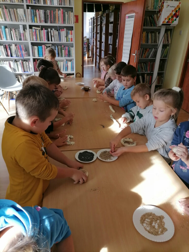 na zdjęciu widoczna jest grupa 15 przedszkolaków stojących przy stołach i wykonujących pieczywo z masy solnej.