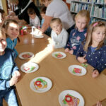 na zdjęciu widoczna jest grupa 13 przedszkolaków stojących przy stolikach i wykonujących papierowe kanapki.