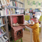 na zdjęciu widoczny jest chłopiec w czapce kucharza, który wkłada do pieca ułożone na łopacie do chleba pieczywo wykonane przez dzieci z masy solnej. W tle regały z książkami i grami planszowymi.