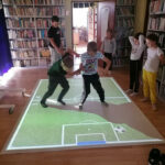 na zdjęciu widoczna jest grupa 7 dzieci (3 dziewczynki i 4 chłopców) biorących udział w zabawach na interaktywnej podłodze FunFloor. W tle regały z książkami.