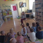 Na zdjęciu widoczna jest grupa przedszkolaków siedzących na matach i słuchających bajki czytanej przez bibliotekarza.
