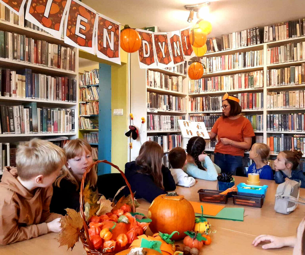 na zdjęciu dzieci siedzące przy stole i słuchające czytającej im bibliotekarki