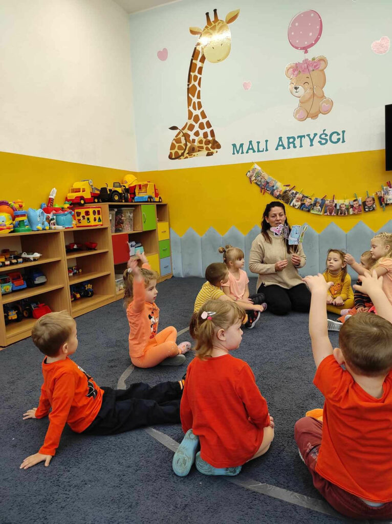 na zdjęciu widać salę przedszkolną, dzieci siedzą w kole na dywanie, bibliotekarz przedstawia treść bajki za pomocą kukiełek