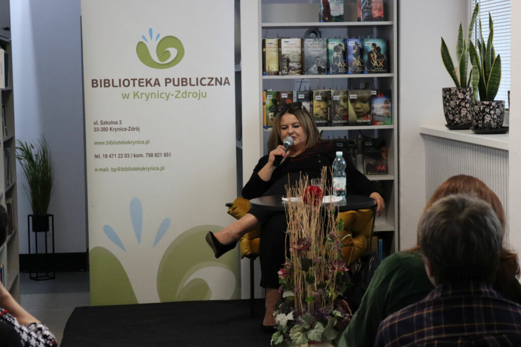 Wnętrze biblioteki. Przy stole okrągłym na fotelu z mikrofonem siedzi autorka spotkania Joanna Jax. Przed nią kwiaty, za nią prezentowane książki.