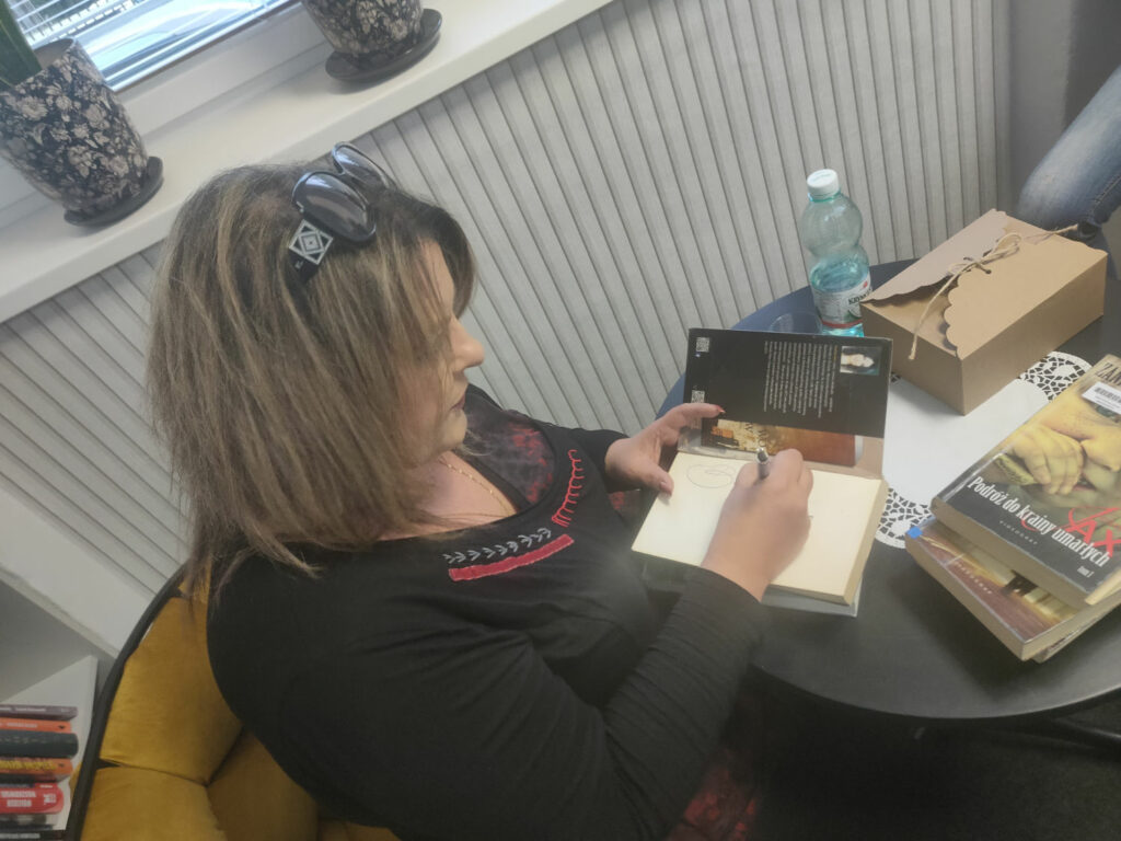Autorka spotkania Joanna Jax siedzi przy stole i podpisuje książki.